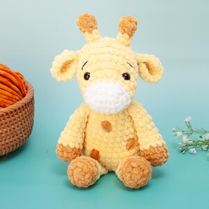 May include: A handmade, plush giraffe toy in shades of yellow and brown. The giraffe has large, floppy ears, small horns, and a white muzzle. The toy is sitting on a light blue surface, with a basket of orange yarn in the background.