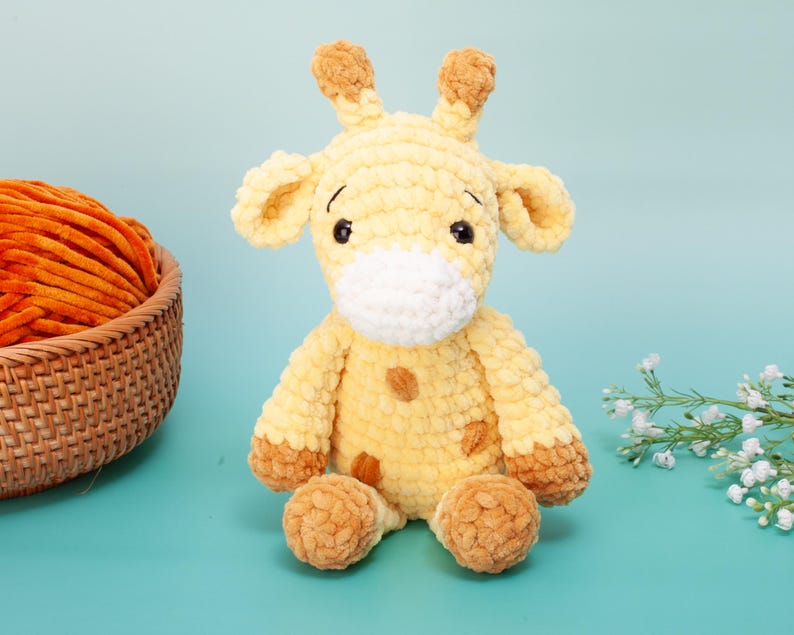 May include: A handmade, plush giraffe toy in shades of yellow and brown. The giraffe has large, floppy ears, small horns, and a white muzzle. The toy is sitting on a light blue surface, with a basket of orange yarn in the background.