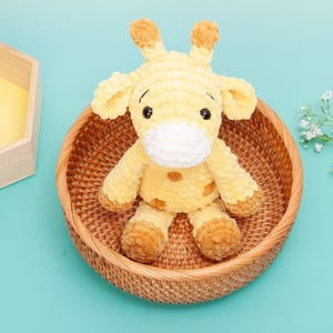 May include: A plush yellow giraffe with brown accents, sitting in a woven basket. The giraffe has large black eyes and a white muzzle. The basket is round and made of natural-colored woven material. The background is a solid light blue.
