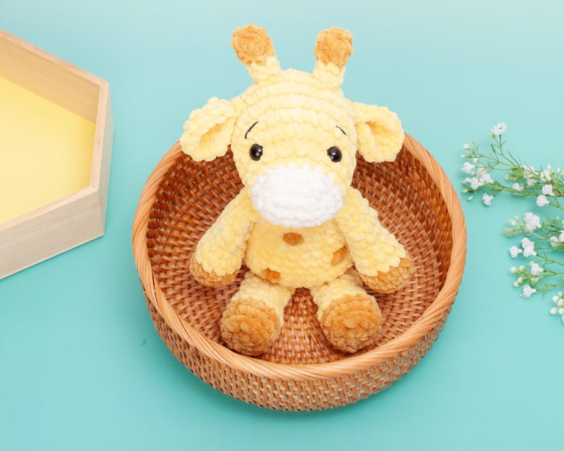 May include: A plush yellow giraffe with brown accents, sitting in a woven basket. The giraffe has large black eyes and a white muzzle. The basket is round and made of natural-colored woven material. The background is a solid light blue.