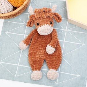May include: A brown crocheted stuffed animal shaped like a cow with white accents on the face and hooves. The cow is lying on a blue and white patterned fabric.