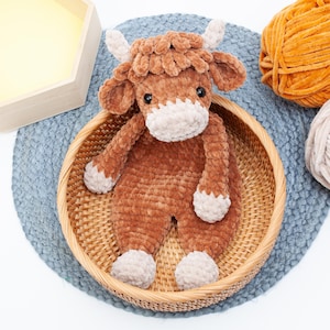 May include: A brown crocheted stuffed animal cow with a white face and hooves. The cow is sitting in a wicker basket on a blue and white rug.