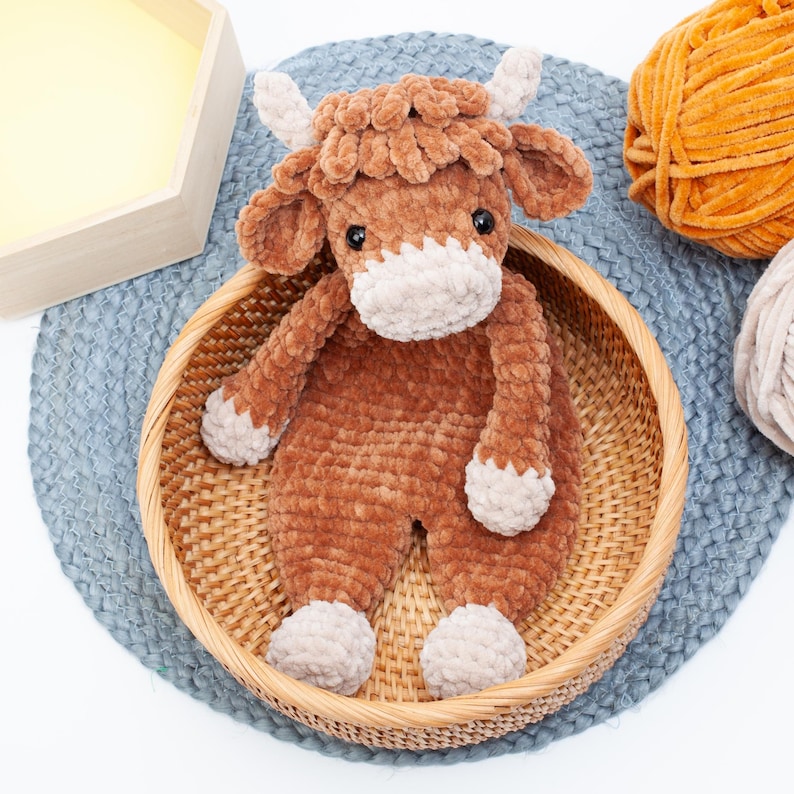 May include: A brown crocheted stuffed animal cow with a white face and hooves. The cow is sitting in a wicker basket on a blue and white rug.