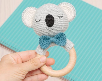 Op de afbeelding: Een gehaakte koala rammelaar met een blauwe strik en een houten ring. De rammelaar is grijs en wit met zwarte ogen.