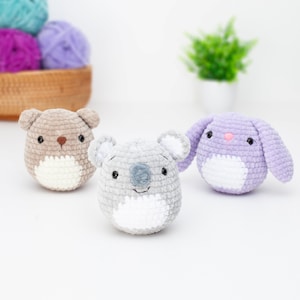 Puede incluir: Tres animales de peluche tejidos a crochet: un oso marrón, un koala gris y un conejito morado. Cada animal tiene una cara blanca con ojos y nariz bordados.