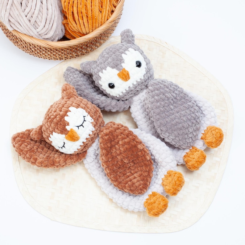 Pu&ograve; includere: Due peluche di gufo all'uncinetto, uno marrone e uno grigio, con gli occhi chiusi e i piedi arancioni. I gufi sono sdraiati su un tappetino intrecciato.