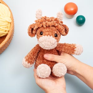 May include: A handmade brown and tan crochet Highland cow plush toy. The plush has a fluffy, textured appearance with small horns and a light tan muzzle and feet. The toy is held in two hands against a light blue background.
