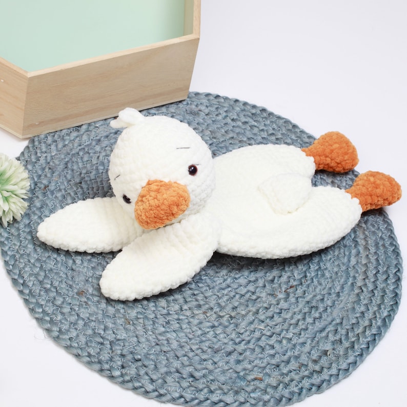 May include: A white crocheted plush toy shaped like a duck with orange beak and feet. The duck is lying on a gray woven rug.