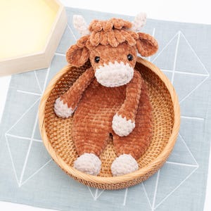 May include: A brown and white crocheted stuffed animal in the shape of a cow. The cow is sitting in a woven wicker basket.