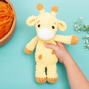 May include: A handmade yellow and brown crochet giraffe toy with a white muzzle and brown spots. The giraffe is being held by a person's hand. A basket of orange yarn is in the background on a turquoise surface.