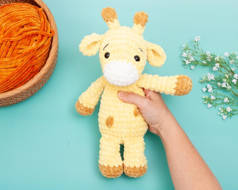 May include: A handmade yellow and brown crochet giraffe toy with a white muzzle and brown spots. The giraffe is being held by a person's hand. A basket of orange yarn is in the background on a turquoise surface.