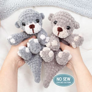 May include: Two handmade crochet teddy bears in shades of gray. The bears have white muzzles, paws, and accents, with dark brown noses and black eyes. The bears are soft and plush, with a textured appearance. The image includes a teal and white oval that says "NO SEW CROCHET PATTERN."