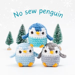 May include: Three crocheted penguin toys in shades of blue and gray, with orange feet and white bellies. The text "No sew penguin" is above the penguins. Small green Christmas trees are in the background.