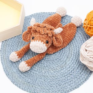 May include: A brown crocheted stuffed animal cow with a white face and hooves. The cow is lying on a blue crocheted mat.
