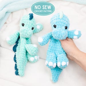 Könnte beinhalten: Zwei gehäkelte Dinosaurier-Spielzeuge in Blau- und Türkistönen. Die Dinosaurier haben kleine schwarze Augen, weiße Akzente an Füßen und Händen und ein strukturiertes, flauschiges Aussehen. Der Text "NO SEW CROCHET PATTERN" befindet sich in einem hellblauen Oval.