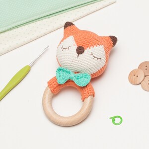 Op de afbeelding: Een gehaakte oranje vossenrammelaar met een groene strik. De rammelaar heeft een houten ring en is een babyspeelgoed.