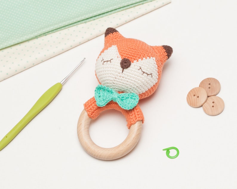 Op de afbeelding: Een gehaakte oranje vossenrammelaar met een groene strik. De rammelaar heeft een houten ring en is een babyspeelgoed.