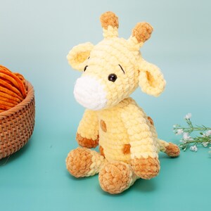 May include: A handmade, plush giraffe toy in shades of yellow and brown. The giraffe has a white muzzle, brown spots, and small black eyes. A basket of orange yarn sits to the left, and a sprig of white flowers is on the right.