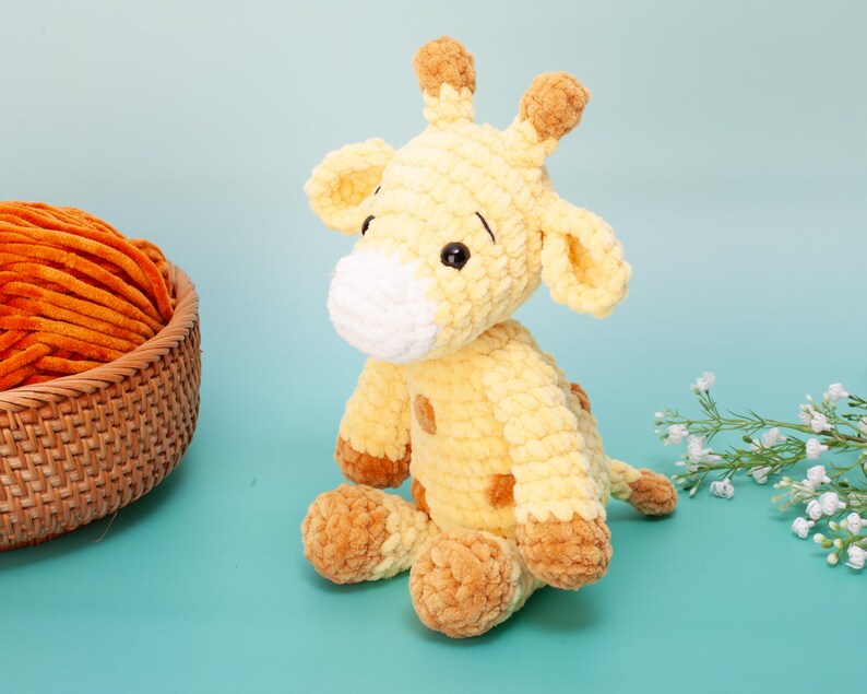 May include: A handmade, plush giraffe toy in shades of yellow and brown. The giraffe has a white muzzle, brown spots, and small black eyes. A basket of orange yarn sits to the left, and a sprig of white flowers is on the right.