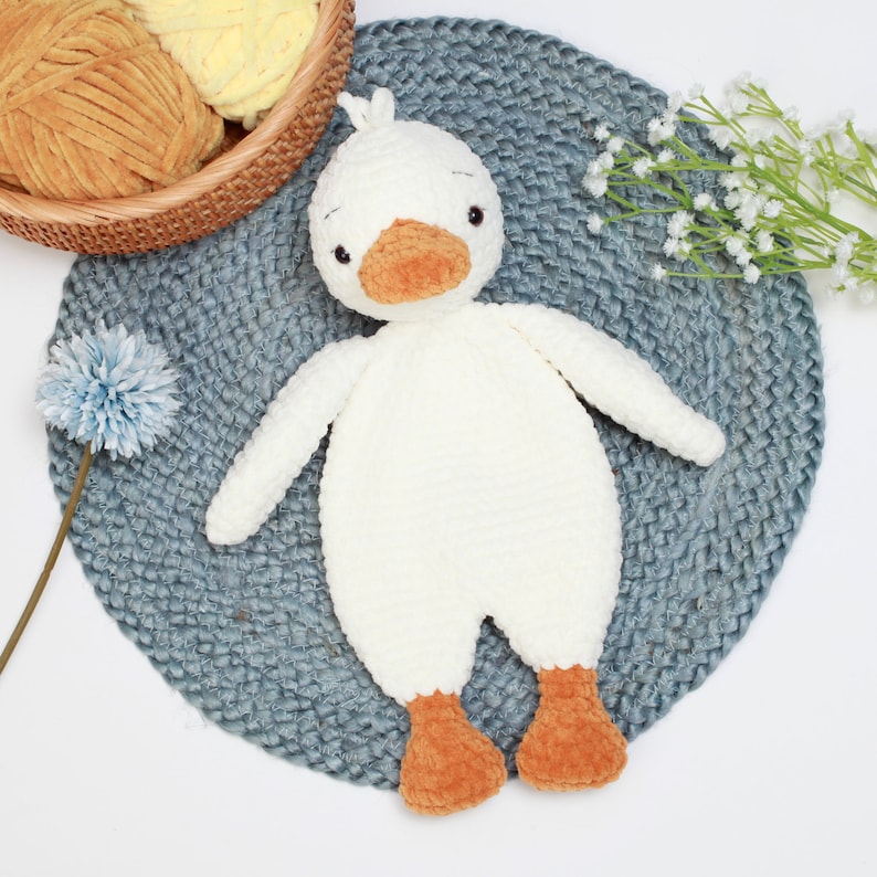 May include: A white crocheted duck plush toy with orange feet and a beak. The toy is lying on a blue and white crocheted mat.