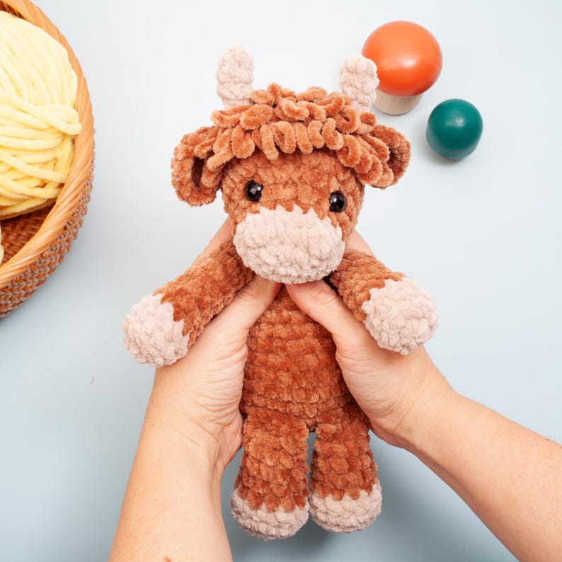 May include: A handmade brown and tan plush Highland cow toy. The plush cow has a fluffy, textured body, small horns, and a light tan muzzle and feet. The toy is held in two hands against a light blue background.