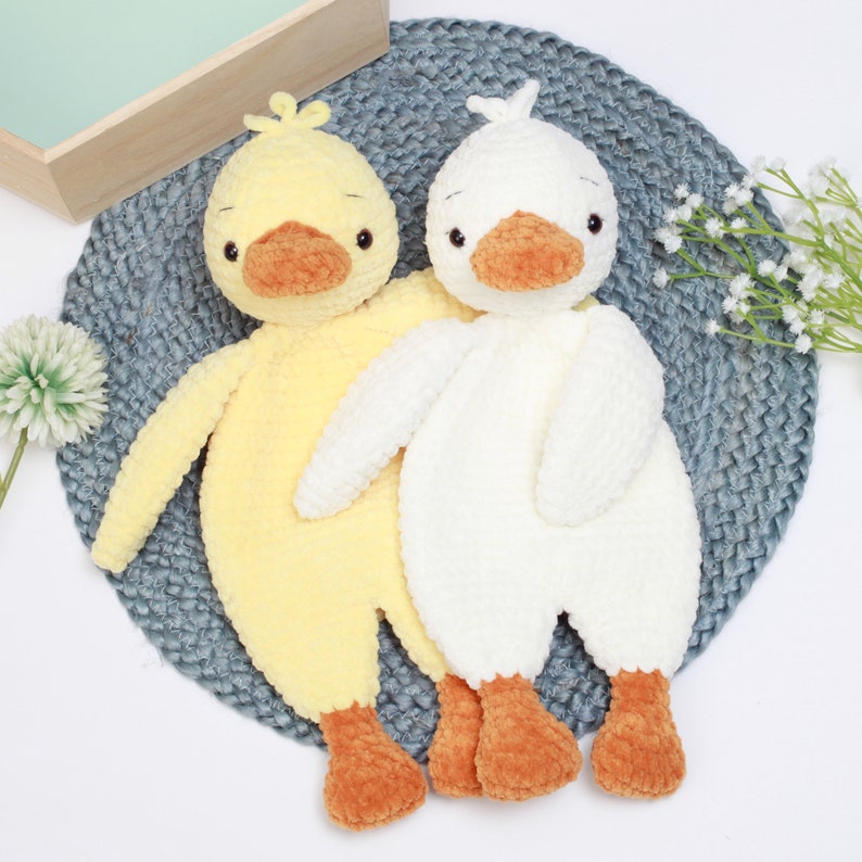 May include: Two crocheted stuffed duck toys, one yellow and one white, with orange beaks and feet. The ducks are sitting on a gray crocheted surface.