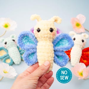 Puede incluir: Una mariposa de peluche hecha a mano en crema y azul, sostenida en una mano. La mariposa tiene ojos grandes y negros y una expresión sonriente. Otras mariposas en menta, blanco y naranja están en el fondo, junto con flores blancas y rosas. Un círculo turquesa con las palabras "NO SEW" está en la parte inferior derecha.
