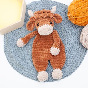 May include: A brown crocheted stuffed animal cow with a white face and hooves. The cow is lying on a blue and grey crocheted rug.