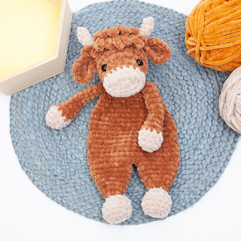May include: A brown crocheted stuffed animal cow with a white face and hooves. The cow is lying on a blue and grey crocheted rug.