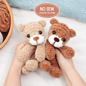 May include: Two handmade crochet teddy bears in light brown and brown. The bears have black eyes, white muzzles, and brown noses. The text "NO SEW CROCHET PATTERN" is in a brown oval above the bears. The bears are soft and plush.