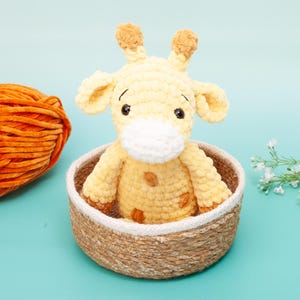 May include: A handmade, yellow crochet giraffe with brown spots and a white muzzle sits in a woven basket. An orange yarn ball is to the left. The background is a light blue.