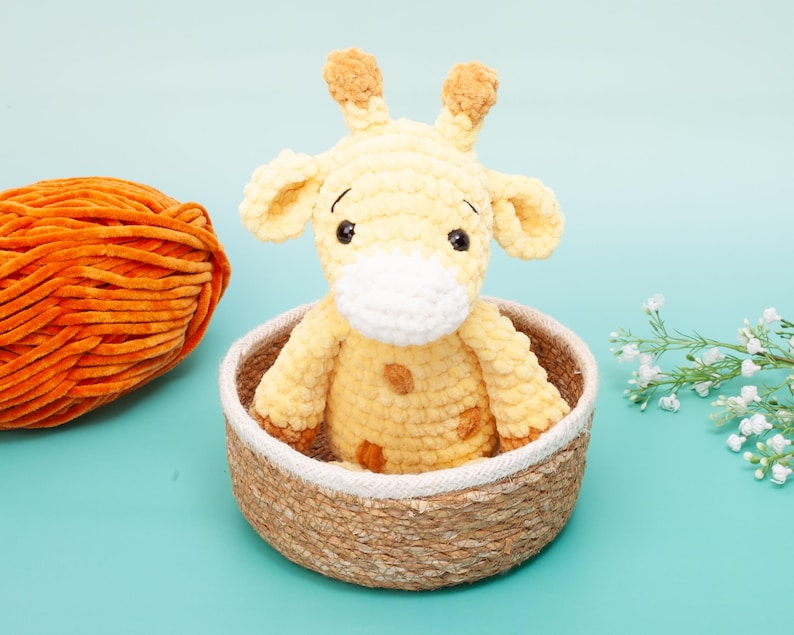 May include: A handmade, yellow crochet giraffe with brown spots and a white muzzle sits in a woven basket. An orange yarn ball is to the left. The background is a light blue.