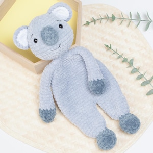 Puede incluir: Un juguete de peluche de koala gris claro hecho a mano, con una nariz y acentos grises. El koala está acostado sobre una estera tejida, con una bandeja de madera en el fondo. El koala tiene pequeños ojos negros y orejas internas blancas.