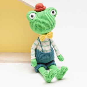 Puede incluir: Una rana de crochet verde con un sombrero rojo, una pajarita amarilla y un mono azul. La rana está sentada sobre una superficie blanca.
