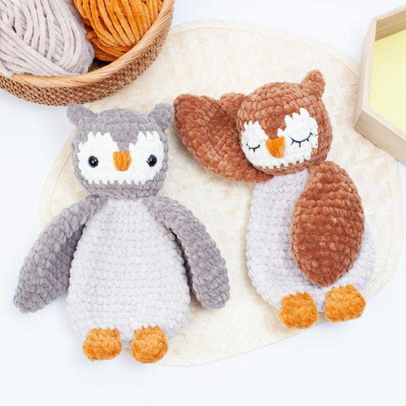 Pu&ograve; includere: Due peluche di gufo all'uncinetto, uno marrone e uno grigio, con gli occhi chiusi e becchi arancioni. I gufi sono seduti su una superficie marrone chiaro.