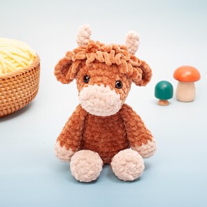 May include: A handmade brown and beige crochet cow plushie with small horns, sitting on a light blue surface. The plushie has a fluffy texture and is accompanied by wooden mushroom decorations and a basket of yellow yarn.