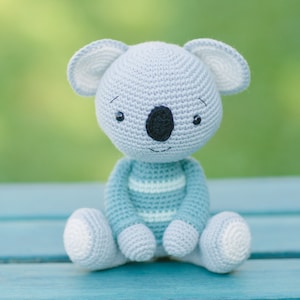 Puede incluir: Un juguete de peluche de koala tejido a crochet con una camisa a rayas azul y blanca. El koala está sentado en una superficie azul.