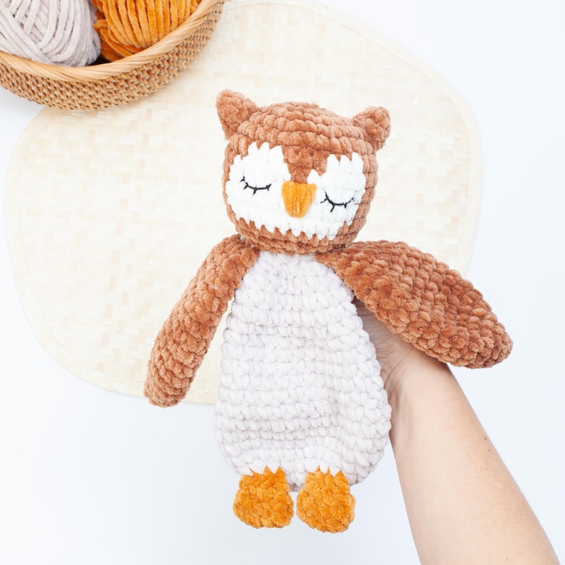 Pu&ograve; includere: Un giocattolo di peluche a forma di gufo lavorato a uncinetto con pelliccia marrone e bianca. Il gufo ha gli occhi chiusi e un becco arancione. Il giocattolo &egrave; tenuto in una mano.