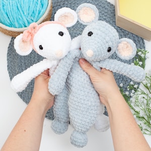 Puede incluir: Dos juguetes de peluche de ratón de crochet, uno blanco con un lazo rosa y otro gris, sostenidos en manos.