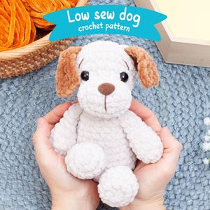 Puede incluir: Un perro blanco de crochet con orejas marrones, sentado en las manos de una persona. El perro tiene una nariz marrón y ojos negros. El fondo es una superficie de crochet azul. El texto "Low sew dog crochet pattern" es visible en la imagen.