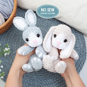 Peut inclure: Deux lapins en peluche, l'un gris clair et l'autre beige, avec des nez roses et des yeux noirs. Les lapins sont faits à la main et duveteux, avec le texte "NO SEW CROCHET PATTERN" dans un ovale bleu.