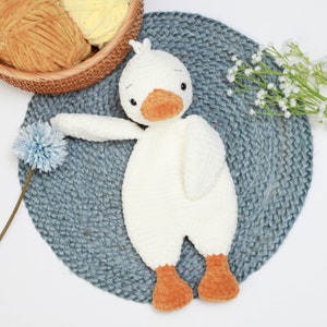 May include: A white crocheted goose toy with orange feet and a beak. The goose is lying on a blue crocheted mat.