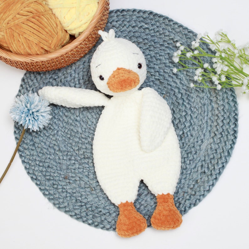 May include: A white crocheted goose toy with orange feet and a beak. The goose is lying on a blue crocheted mat.