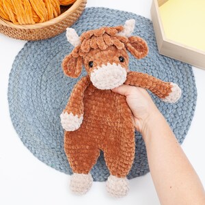 May include: A brown crocheted stuffed animal cow with a white face and hooves. The cow is sitting on a blue crocheted mat.