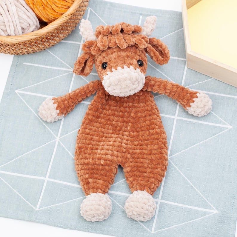 May include: A brown crocheted stuffed animal shaped like a cow with a white face and hooves. The cow is lying on a blue and white patterned fabric.