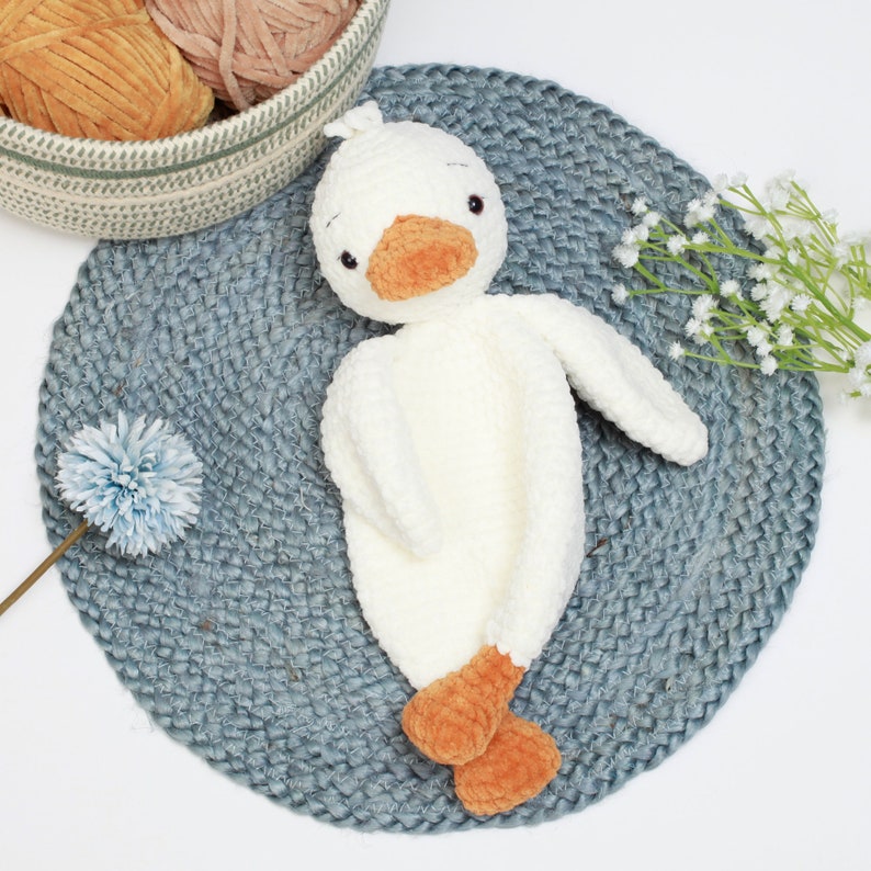 May include: A white crocheted duck plush toy with orange feet and a beak. The duck is lying on a blue and white woven rug.