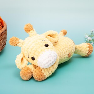 May include: A handmade, yellow and brown crochet giraffe toy with black safety eyes and a white muzzle. The plush giraffe is lying down on a light blue surface. The giraffe's ears, hooves, and spots are brown. A basket of orange yarn is in the background.