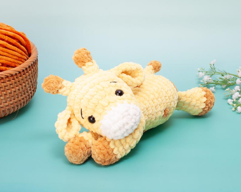 May include: A handmade, yellow and brown crochet giraffe toy with black safety eyes and a white muzzle. The plush giraffe is lying down on a light blue surface. The giraffe's ears, hooves, and spots are brown. A basket of orange yarn is in the background.