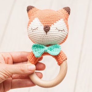 Op de afbeelding: Een gehaakt vosje rammelaar met een houten ring en een groene strik. Het speelgoed is oranje en wit met bruine oren en een bruine neus.