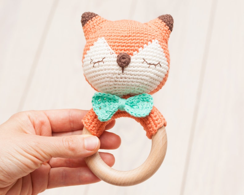Op de afbeelding: Een gehaakt vosje rammelaar met een houten ring en een groene strik. Het speelgoed is oranje en wit met bruine oren en een bruine neus.